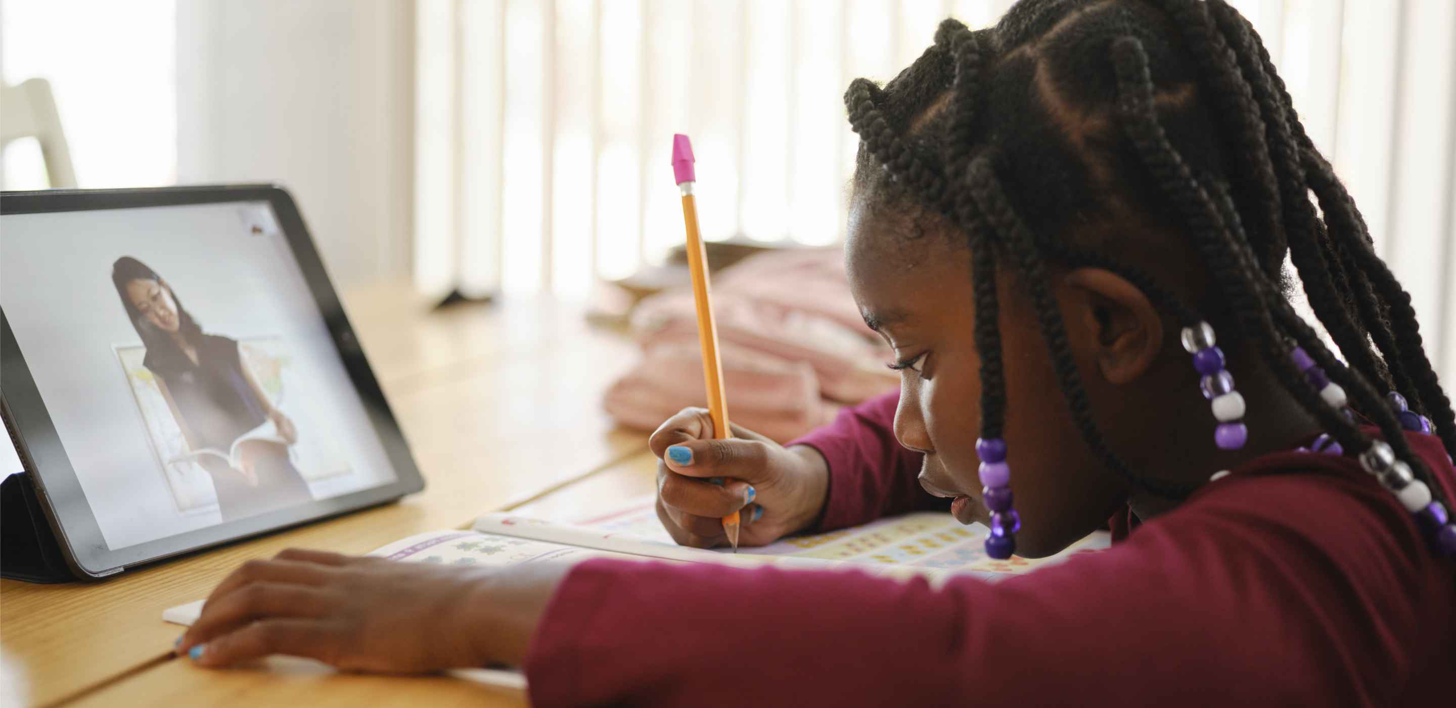 child sitting and writing at desk with active tablet device facing her  