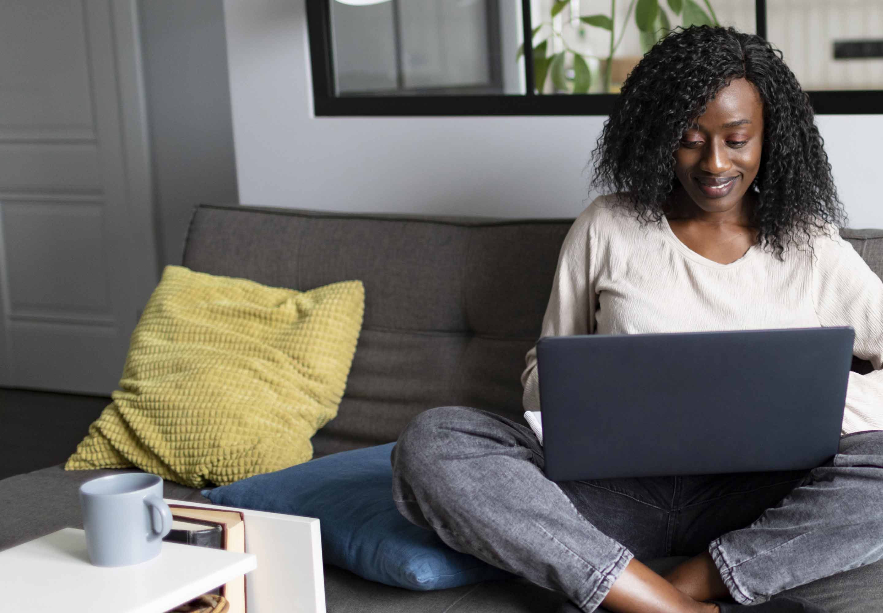 Smiling young black woman sitting legs crossed on couch with laptop on lap
